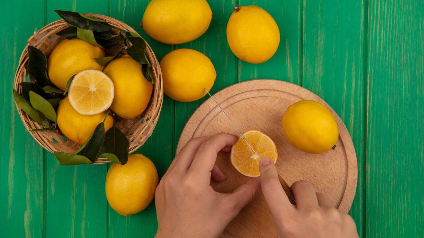 top-view-female-hands-cutting-lemon-wooden-kitchen-board-with-knife-with-lemons-bucket-green-wooden-wall_141793-76020