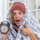 unhealthy-young-woman-hat-wrapped-blanket-holding-alarm-clock-surprsied-sitting-couch-light-living-room_141793-101912