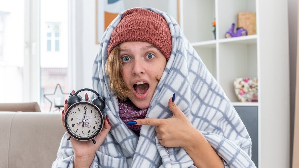 unhealthy-young-woman-hat-wrapped-blanket-holding-alarm-clock-surprsied-sitting-couch-light-living-room_141793-101912